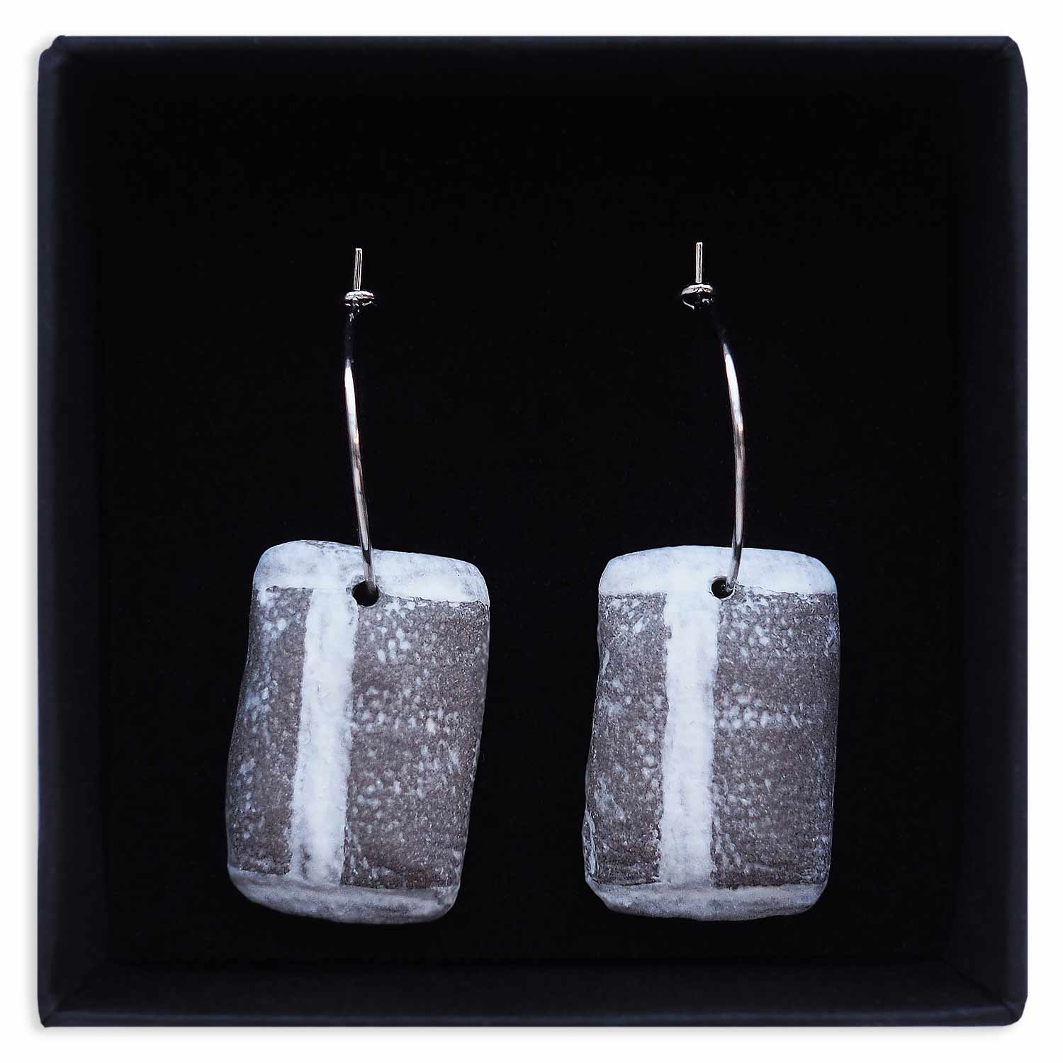 Ett par örhängen ligger i en svart, kvadratisk smyckesask. Örhängena är silverfärgade creoler med snusprillor av keramik som berlocker.