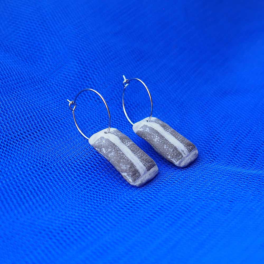 Ett par örhängen ligger på blått tylltyg som bakgrund. Örhängena är silverfärgade creoler med snusprillor av keramik som berlocker.